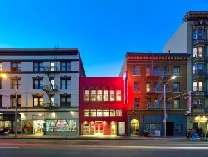 A two-story magenta building is sandwiched between two larger apartment buildings at dusk.
