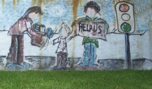 A large graffiti'd mural, painted in a cartoonish style, showing an apparently homeless family of three standing near a traffic light. The mother is handing a box or carton of stuff to the child, whose other hand is reaching to the father figure. He is holding up a sign that says "Help us." Facial features were not drawn on these figures, giving them a universal quality.