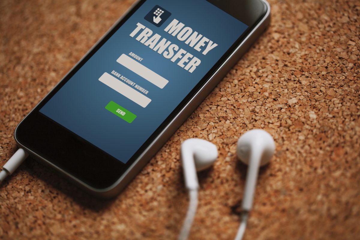 Stock image of mobile phone lying on a corkboard with screen reading "Money Transfer" and fields for amount and account number (not filled in). A pair of white earbuds is connected to the phone and also lying on the cork.