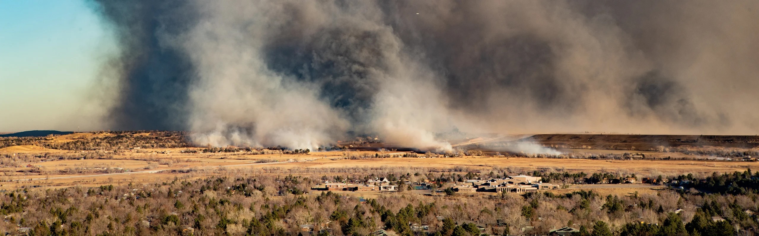 Distant view of wildfire in Colorado. In the middle distance are houses and buildings. Beyond them, on the far side of an open landscape of grasses, are more settled areas. Some of the buildings are burning. Over the distant ground, thick dark smoke covers the right-hand seven-eighths of the image.At far left is a bit of blue sky.