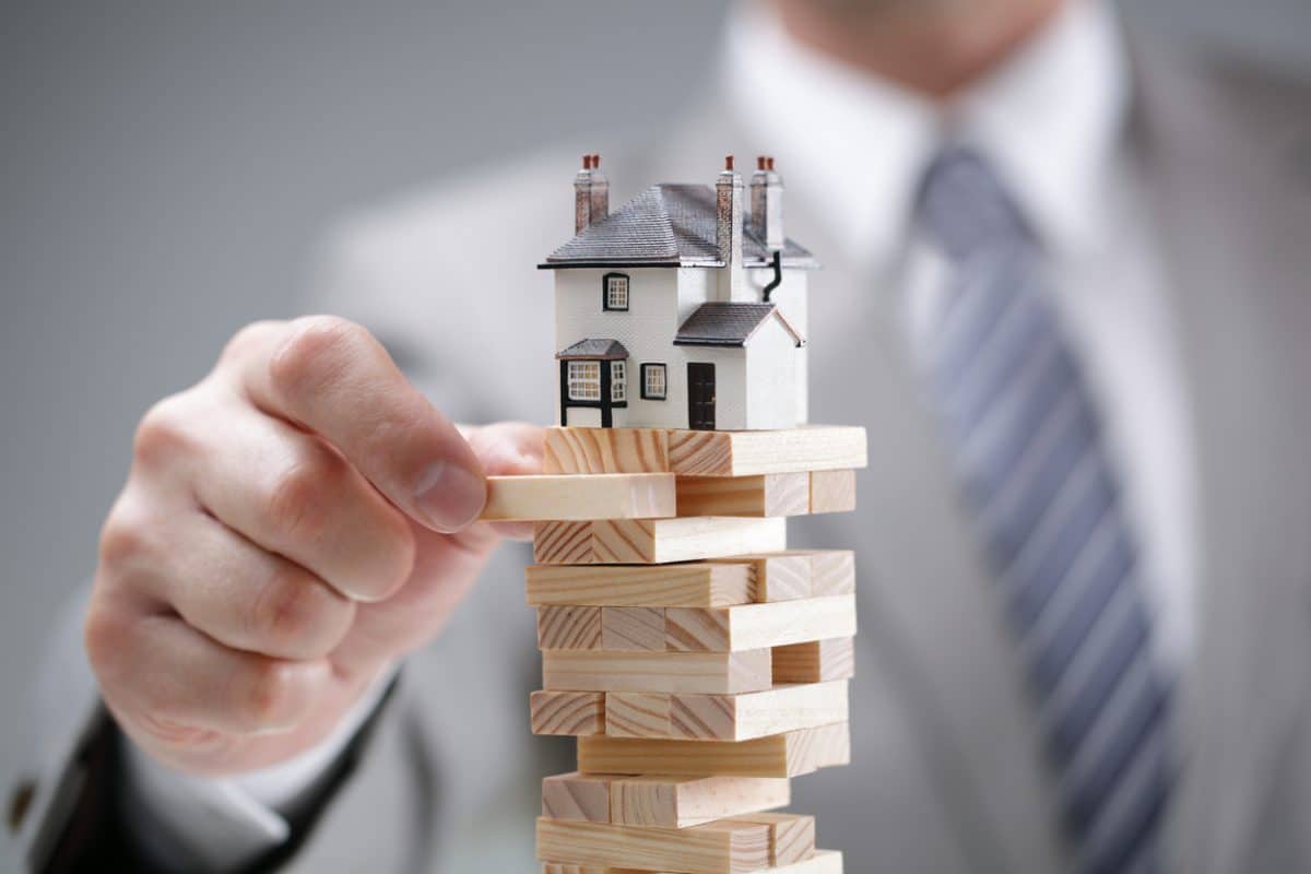 In focus in foreground is a tower of small blocks atop which is a tiny model of a white house with gray pitched roof. Behind the house is a white man in a business suit, out of focus except for his hand, which is holding one of the blocks supporting the house, as if to pull it away.