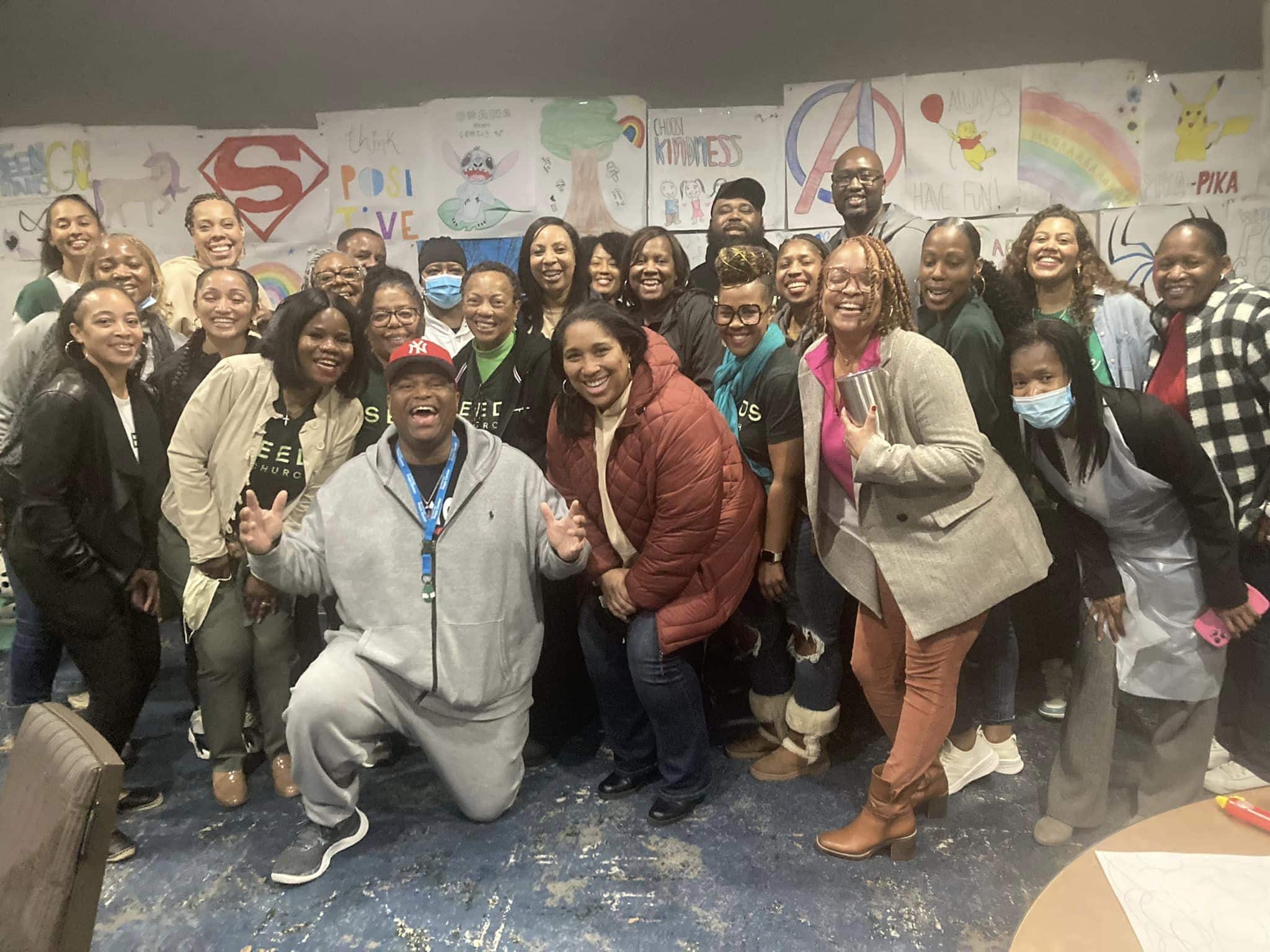 About 25 people in a range of skin tones pose for a photo, all smiling. Most are in cool-weather jackets and two wear surgical masks. A woman at right is wearing a plastic apron. Behind the group are hand-done posters with messages of kindness and love.