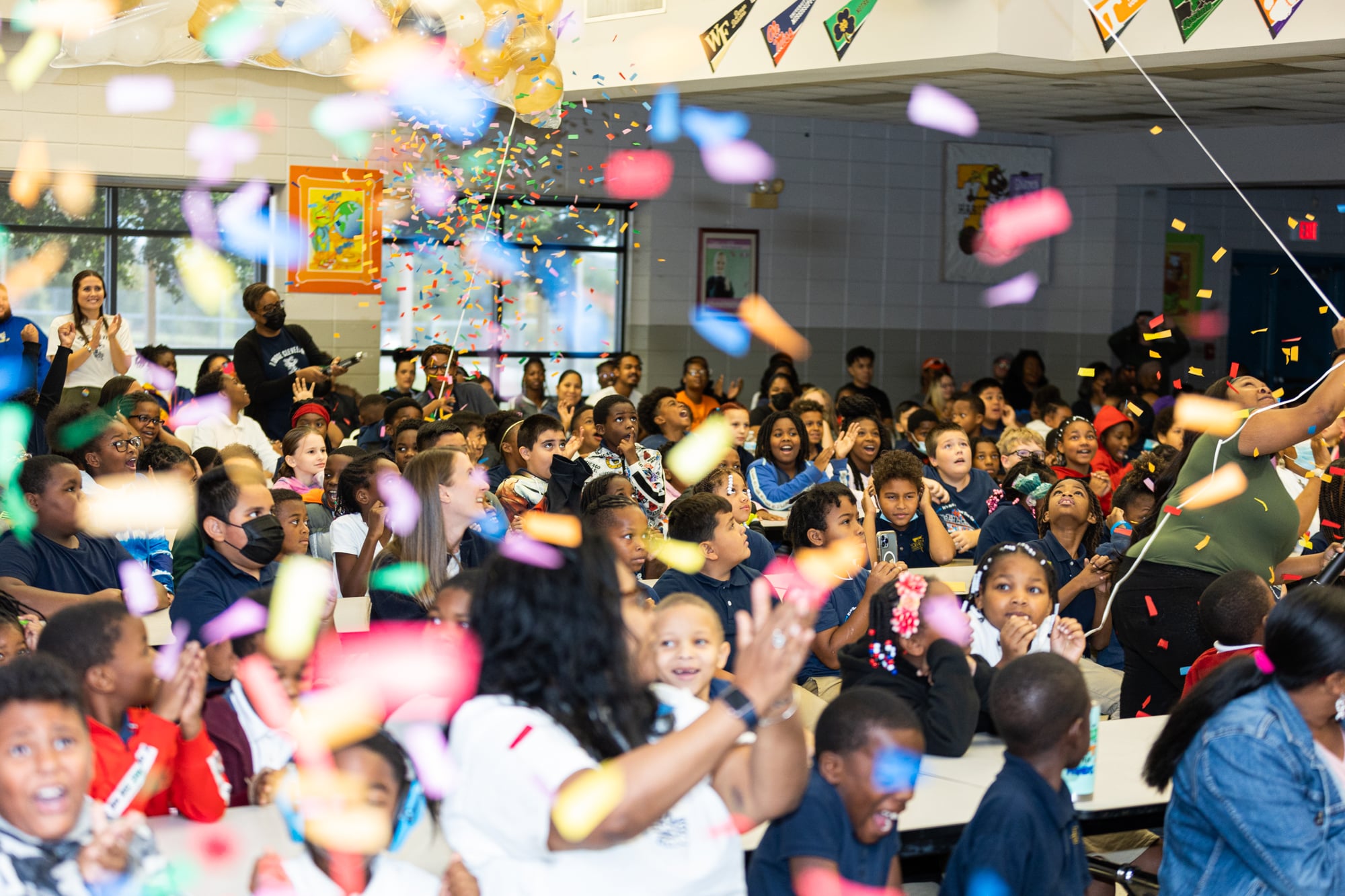A school cafeteria or other large room crowded with elementary school kids of varying skin tones. The air above them is filled with confetti and many are clapping. Open mouths indicate cheering or yelling. At the back of the room are three adults, two clapping and one operating the confetti blower.