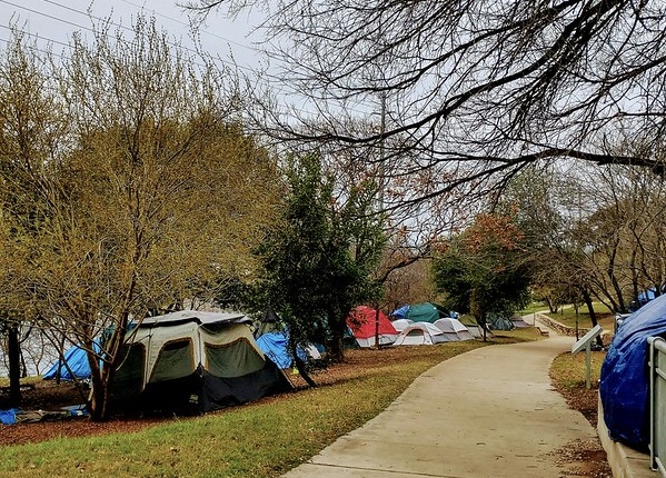 A number of tents pitched on the buffer zone between the road and sidewalk. At least nine tents are discernible among the trees and shrubs. Photo taken in early spring; the trees are just in bud.