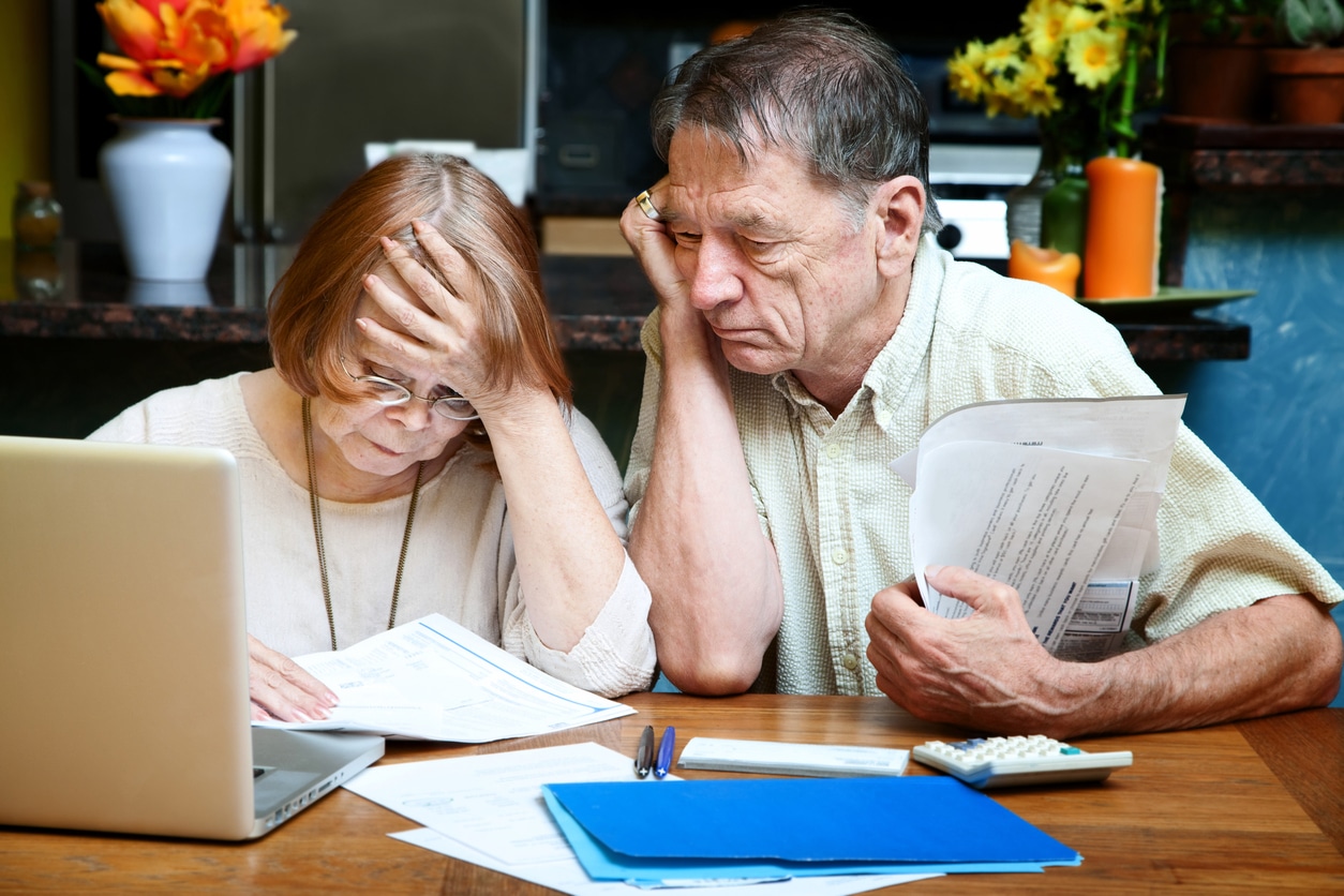 An older white couple sits side by side at a table covered with paper, folders, calculator, pens, and laptop. They're looking at a document with apparent worry or anxiety.