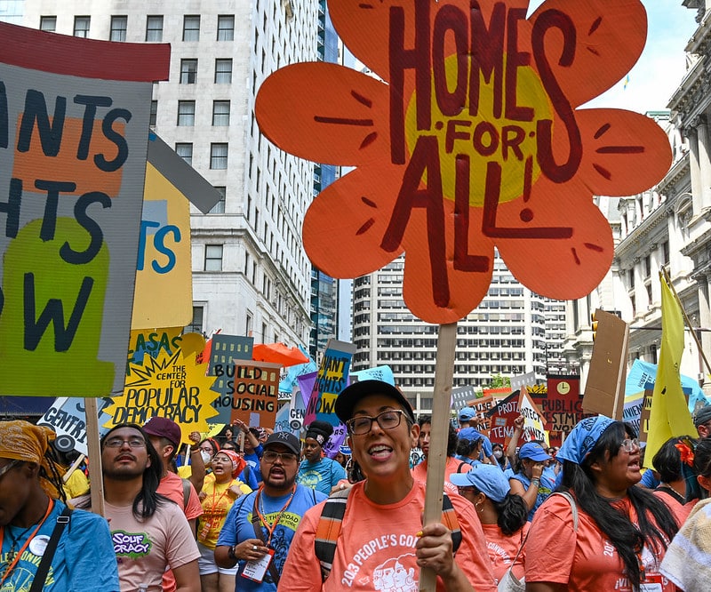 A crowd of marchers in a city street of high-rises and older buildings, most carrying colorful hand-lettered signs advocating housing for all. In the foreground, a person in an orange T-shirt carries a large daisy-shaped sign that reads "Homes for All"