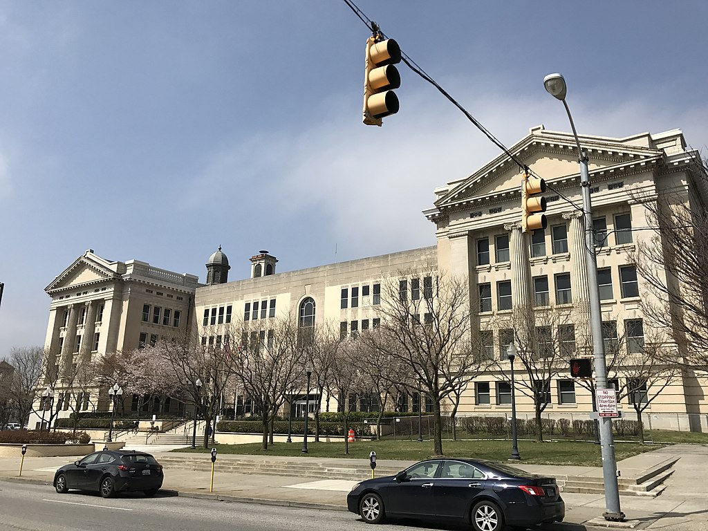 Large imposing building with two peaked-roof five story sections joined by a lower section with a flat roof. The two taller parts have Greek columns. Trees in front are beginning to bud; two cars are parked at meters along the curb.