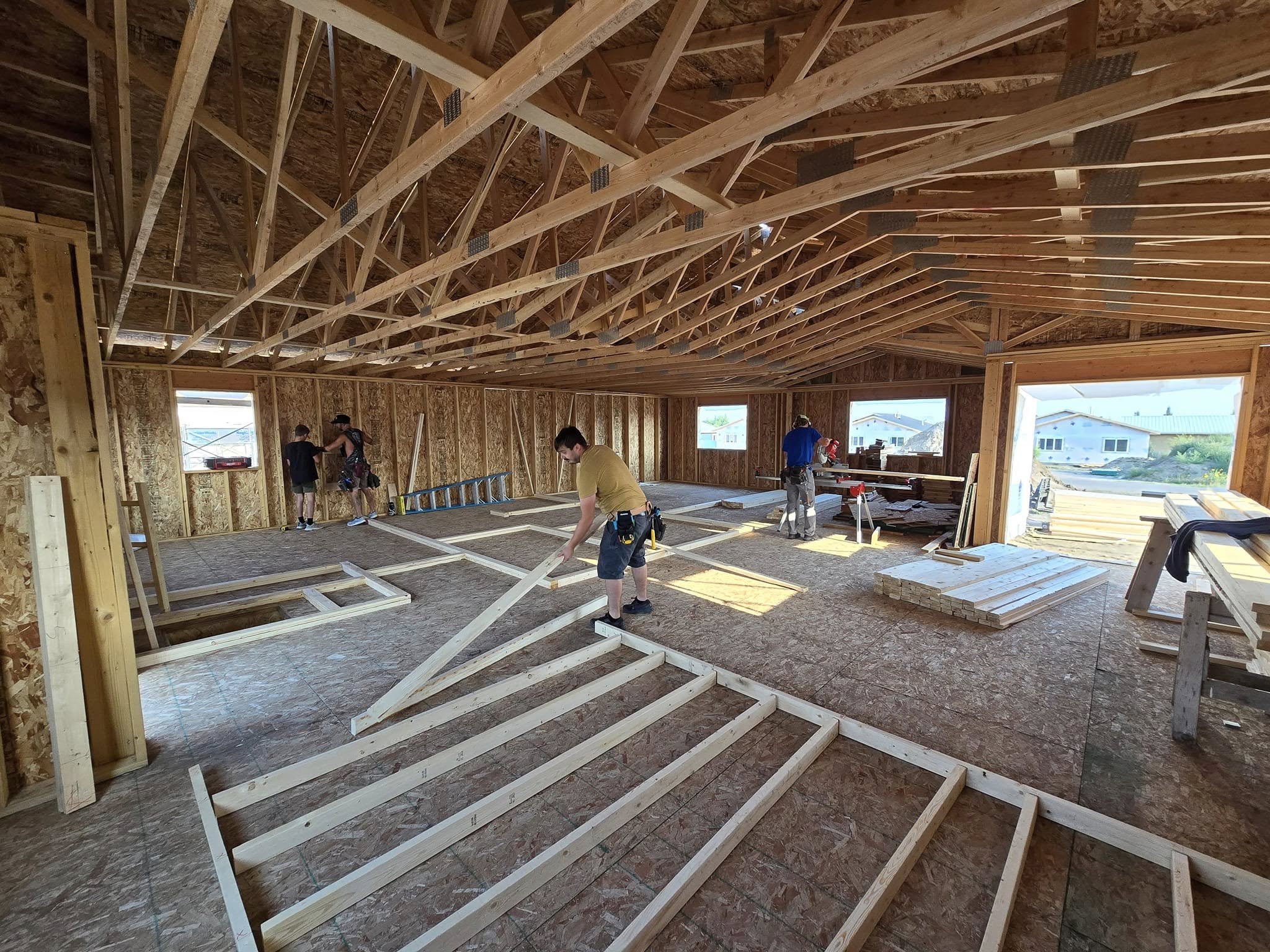 Inside a framed peaked-roof house, four people are working. One is laying two-by-fours on the floor to frame a wall; one is using a table saw, and two are standing at the far wall looking at something out of camera view. Out the large doorway (which has no door yet) can be seen a house across the street, and piles of lumber.