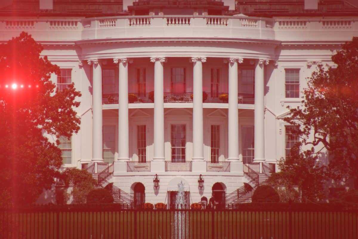 To represent the Trump administration: The portico of the White House overlooking the South Lawn. An imposing building with columns marking a two-story portico and a balcony on the second floor. Angled staircases on either side lead up to the first floor. On the lawn, trees in leaf frame the portico on either side. The photo has an eerie red tint.